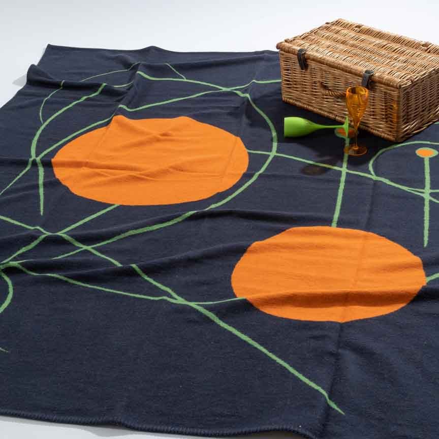 Black picnic blanket with orange circles and green lines, featuring a wicker picnic basket and glasses.