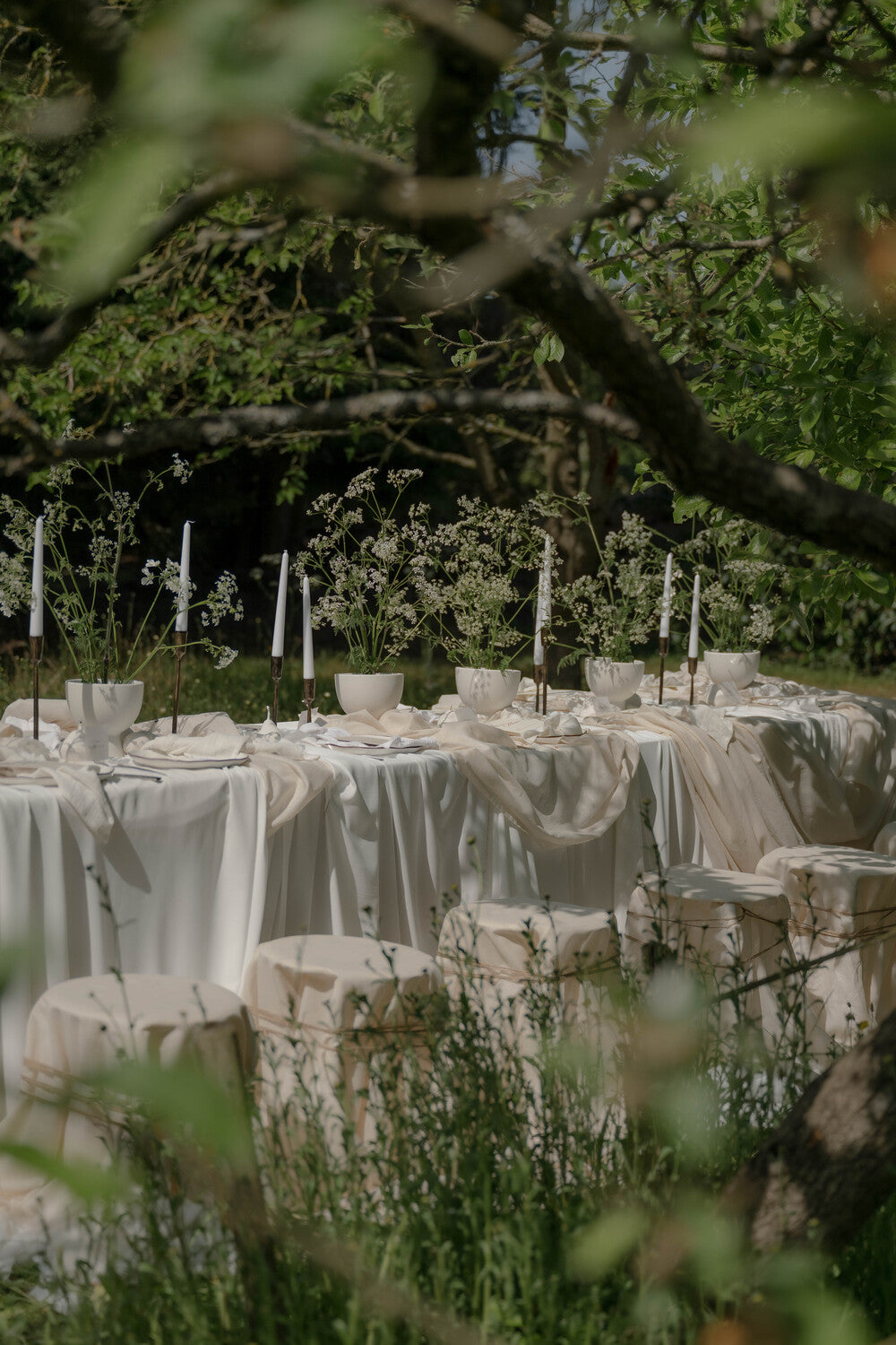 Outdoor Wedding Table Rustic and elegant with white natural linens drapped across, candle holders with white taper candles, surrounded by trees and greenery.