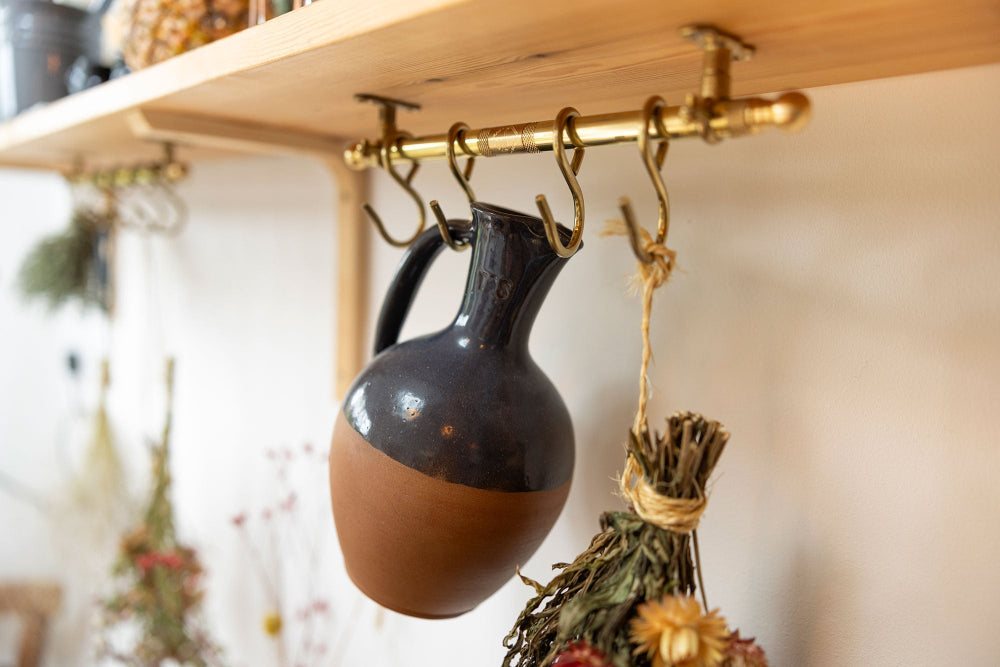 A rustic two-toned ceramic jug with a glossy black upper half and terracotta brown lower half, hanging from brass wall hooks on a wooden shelf.