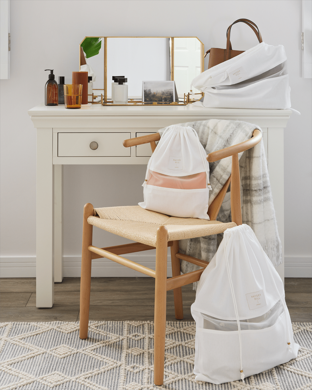 White drawstring mesh garment bags, including one with a peach-colored inner pouch, displayed on a wooden chair next to a white vanity table in a bright, modern room.