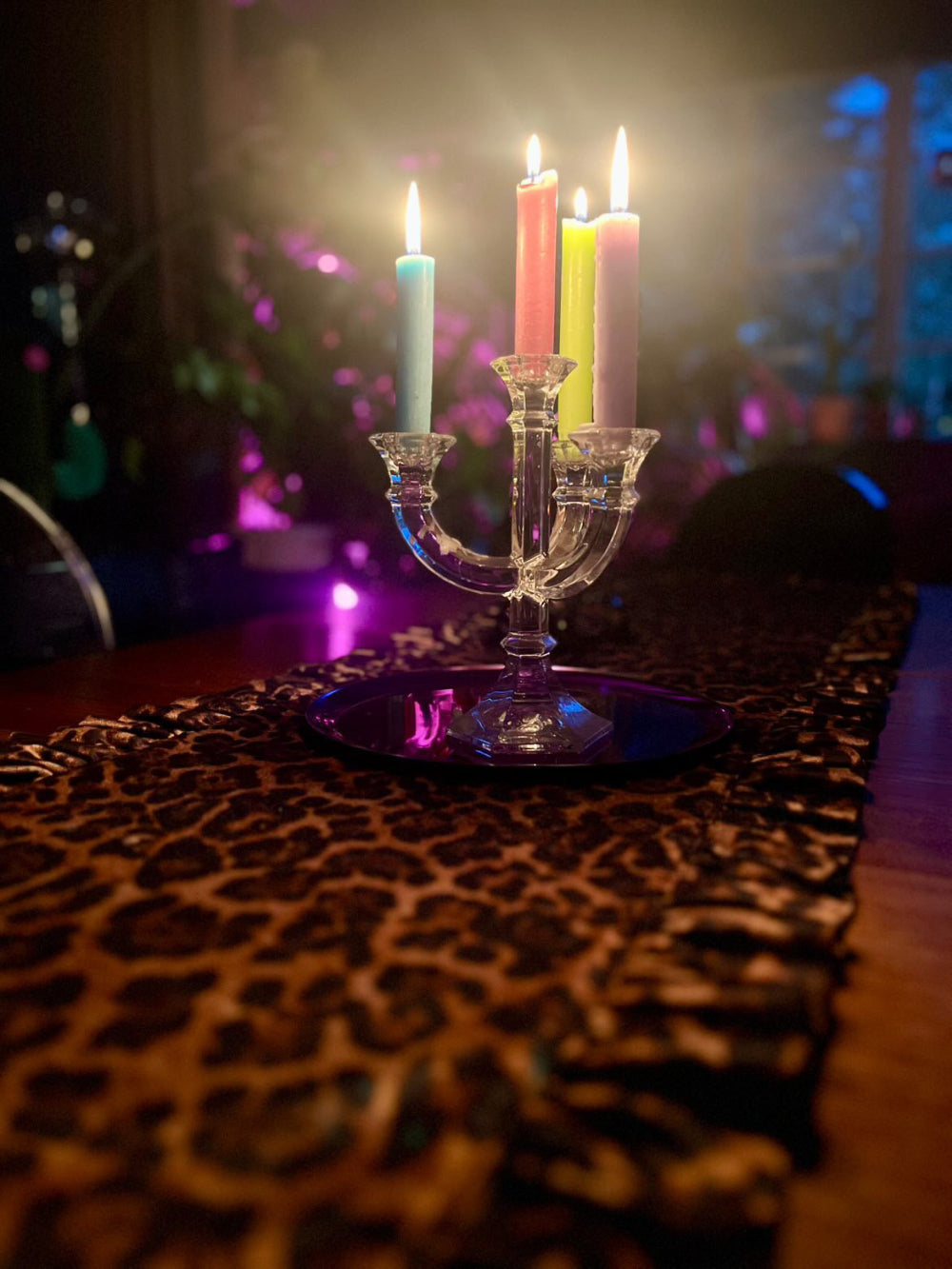 A clear glass three-arm candelabra holding three lit taper candles in pastel blue, pink, and yellow, placed on a dark tray atop a leopard-print table runner in a dimly lit room with colorful ambient lighting.