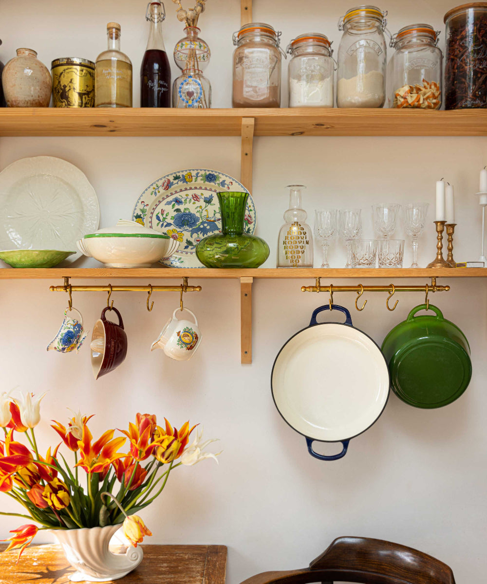 A styled kitchen shelf scene with wooden shelves holding glass jars, dishes, cups, and cookware, plus brass hooks hanging mugs, pots, and pans against a white wall. A vase of orange flowers sits on a table in the foreground.