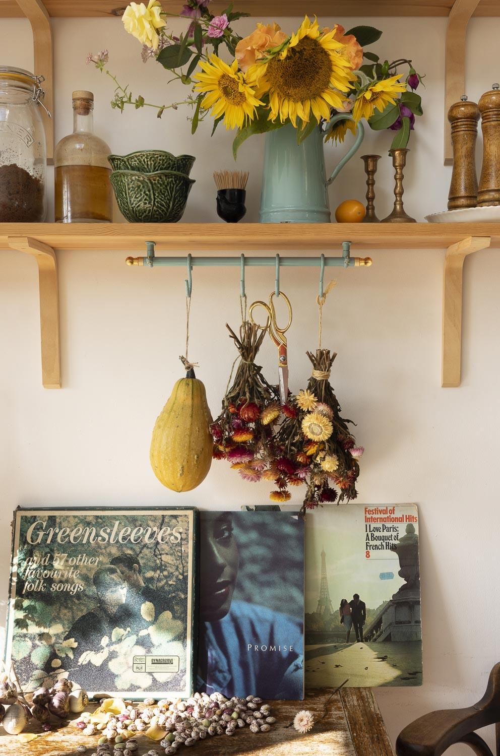 A styled interior shelf scene with wooden shelves, a turquoise pitcher of sunflowers, hanging dried floral bundles on a teal rack, a yellow squash, books, and decorative objects against a white wall.