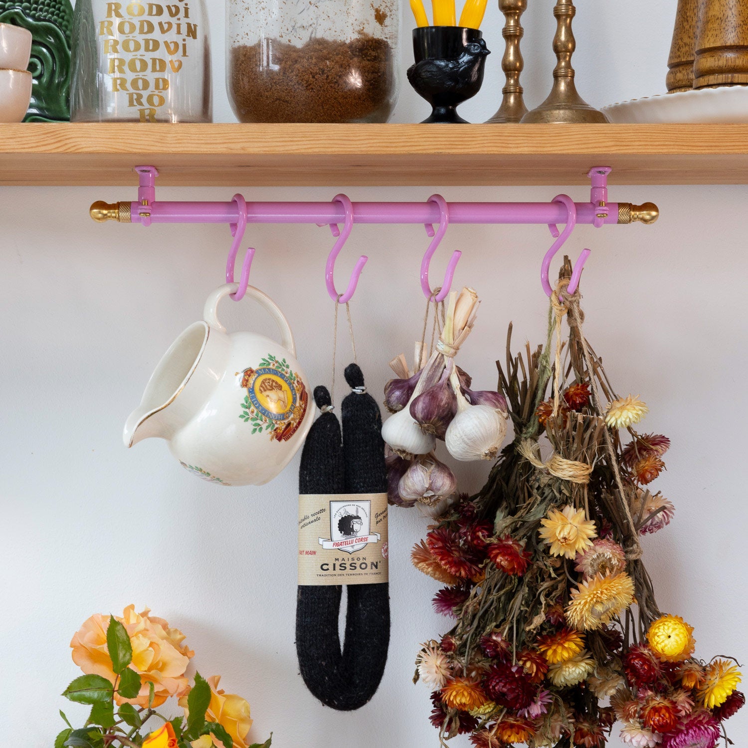 A pink wall-mounted hook rack with brass-toned end caps hangs on a wooden shelf. Several kitchen items are suspended from the hooks, including a white ceramic pitcher, black knit socks, garlic, dried flowers, and other home decor.