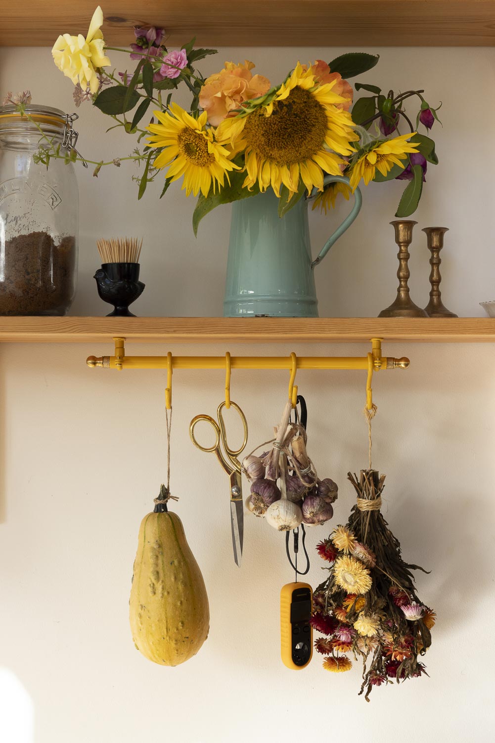 A rustic wall-mounted brass hook rack with yellow hanging cords holds kitchen and garden items, including a yellow squash, scissors, garlic, dried herbs, and a small black tool. Above the rack, a wooden shelf holds a mint green pitcher filled with sunflowers and other flowers, alongside a glass jar and brass candlesticks.