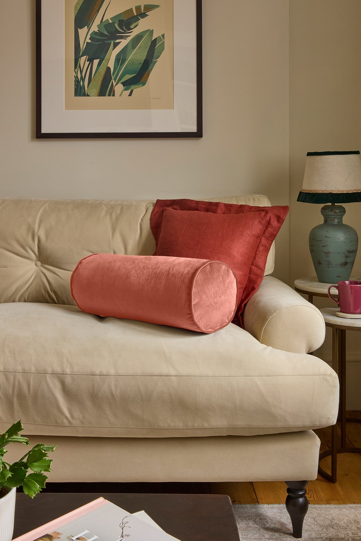 A styled living room scene featuring a cream-colored sofa with two decorative pillows: a cylindrical dusty rose pillow in front and a rectangular rust-red pillow behind it. A framed botanical print hangs on the beige wall above, and a blue-green table lamp sits on a side table to the right.