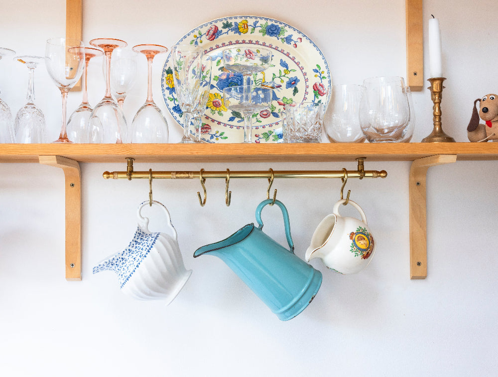 A brass wall-mounted mug rack hanging from a wooden shelf, holding three ceramic mugs. The shelf is styled with a floral serving platter, clear drinking glasses, and a white candle against a white wall.