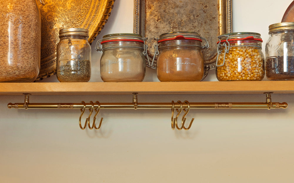 A brass wall-mounted rod with three hanging hooks beneath a wooden shelf holding several glass jars filled with spices or grains against a warm beige wall.