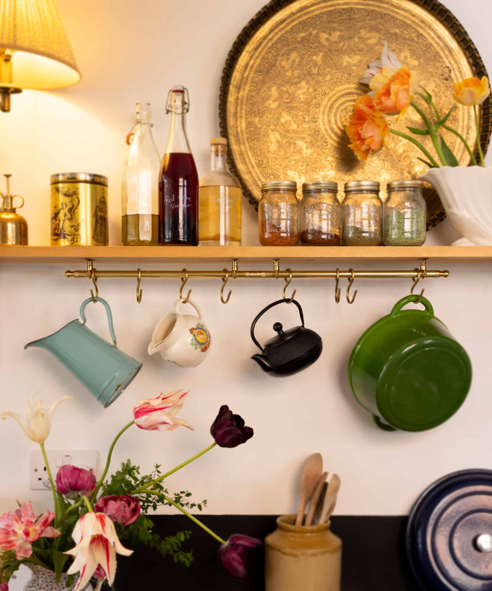 A styled kitchen shelf and wall hook rack with hanging cookware and glass bottles, including a teal pitcher, white teapot, black kettle, green pot, flowers, and decorative jars against a warm cream wall.
