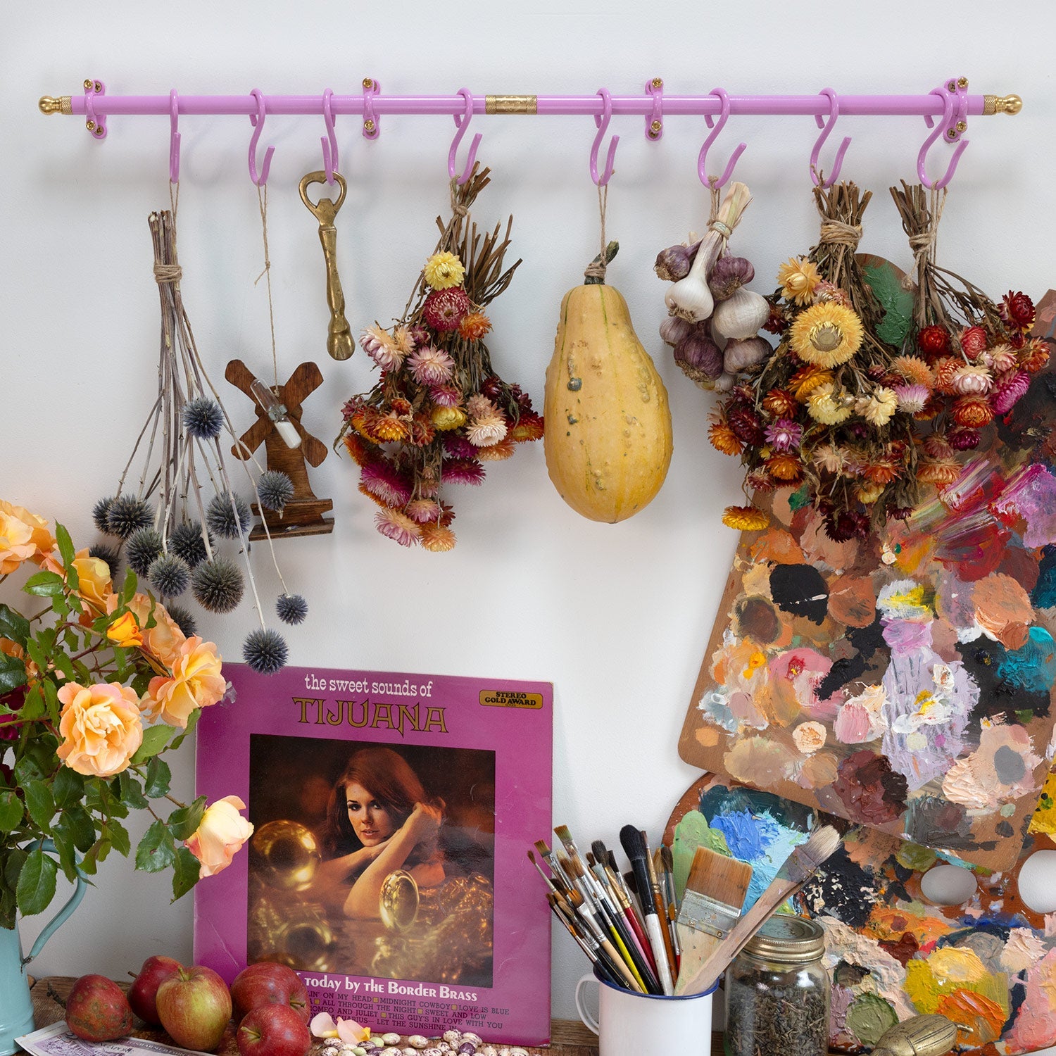 A styled interior scene with a pink wall-mounted rod holding dried flower bundles, a wooden cross, a vintage key, a yellow squash, garlic, and other decorative objects. A book titled 'The Great Gatsby of Tijuana' lies on the floor, alongside paintbrushes, jars, apples, and a colorful paint palette against a white wall.