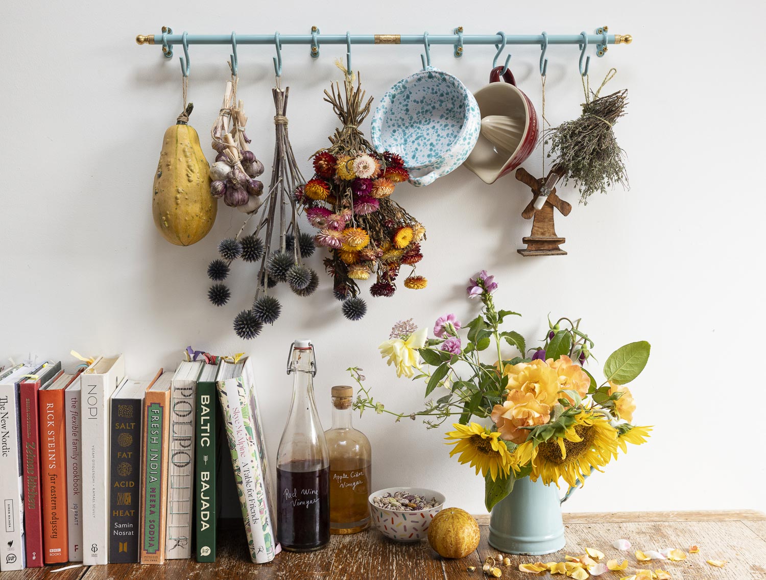 A rustic wall-mounted teal rack with hooks displays dried flowers, a blue speckled bowl, mugs, herbs, and a small wooden bird figurine. Below, a wooden table holds a vase of yellow sunflowers, books, glass bottles, and a small bowl on a white wall.