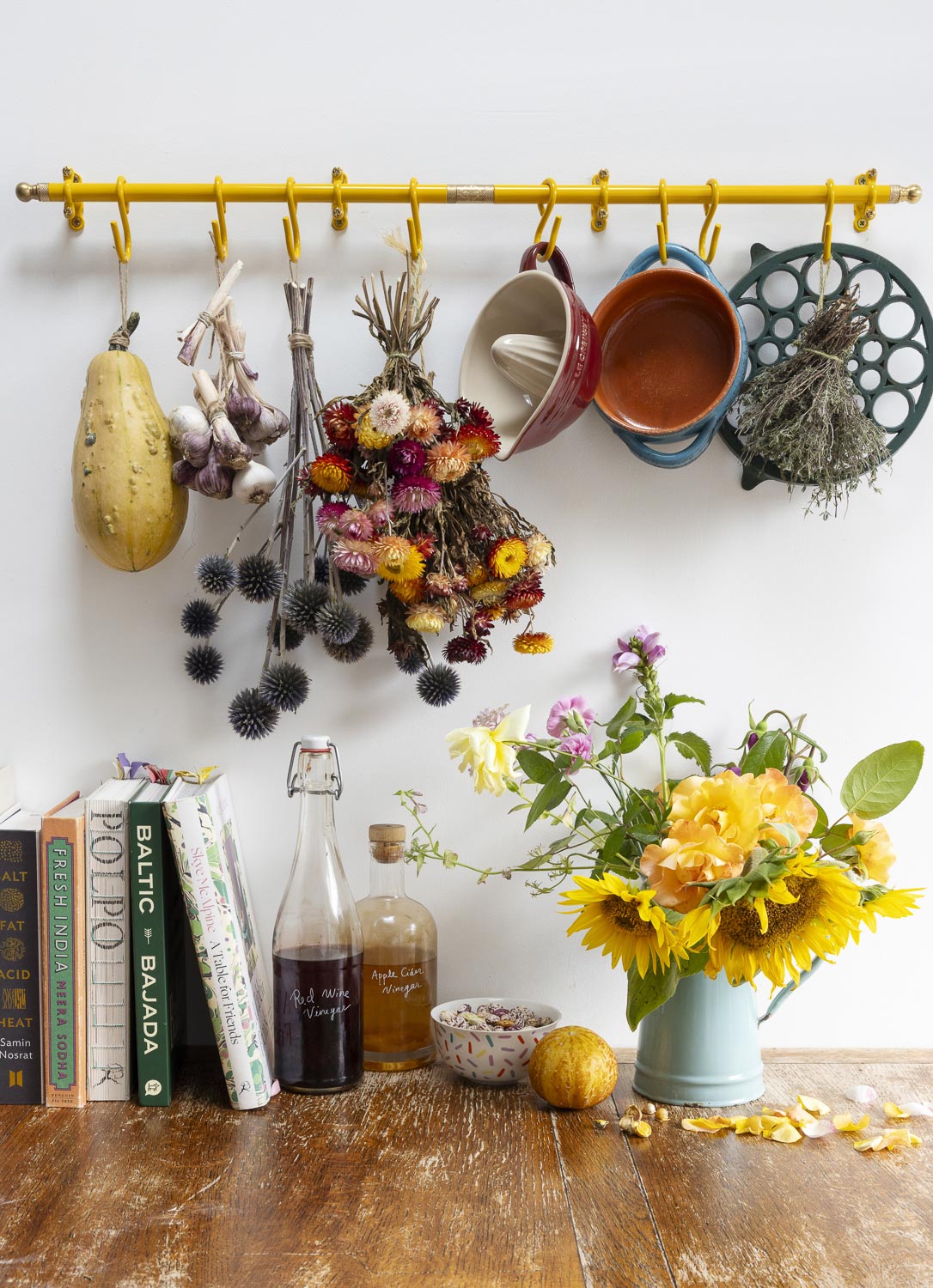 A rustic kitchen wall display featuring a bright yellow pegboard rack holding colorful ceramic mugs, dried flower bouquets, herbs, and a yellow squash. Below the rack, a wooden countertop holds books, glass bottles, a bowl, and a light blue tin filled with sunflowers.