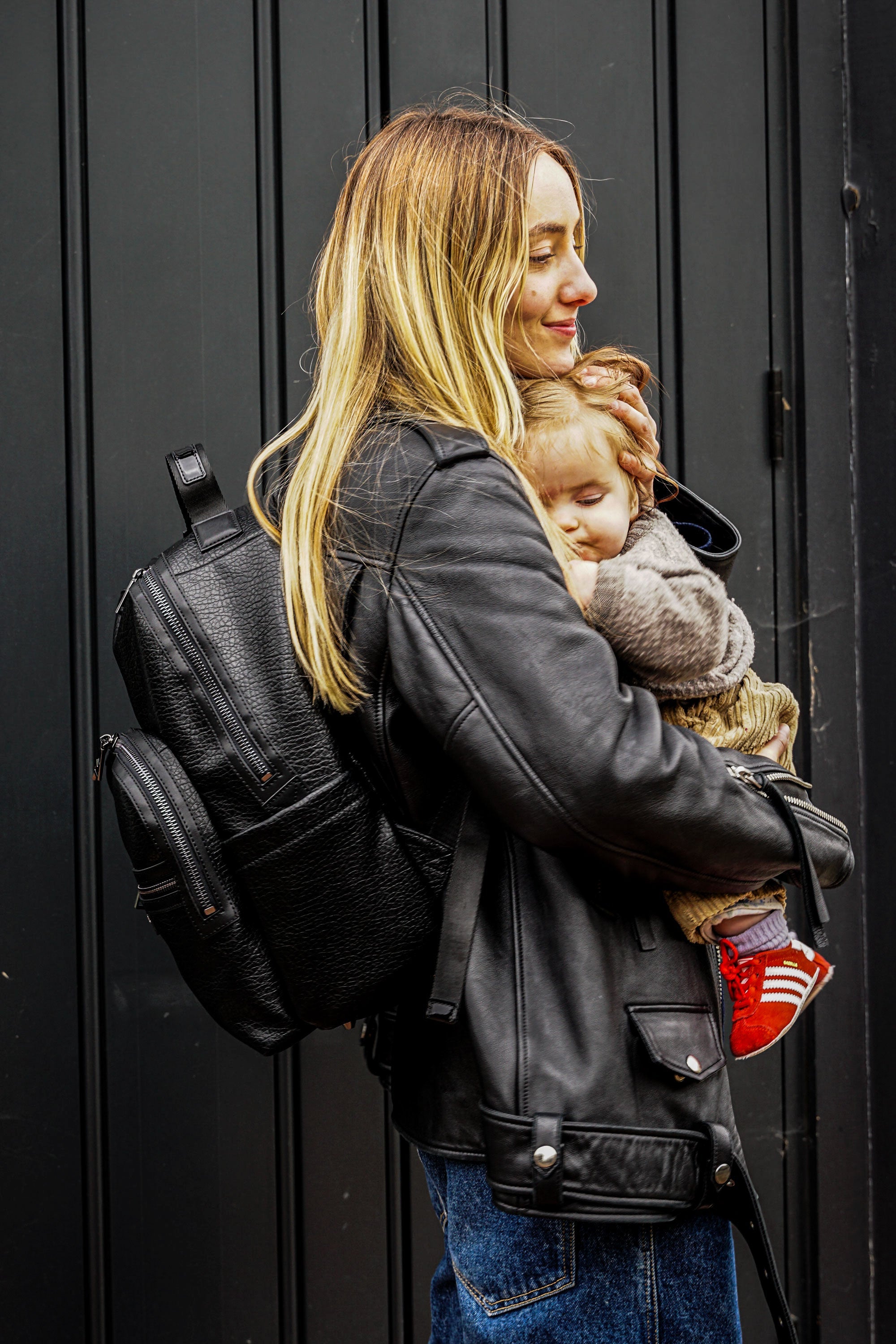 Woman carrying a child and a black backpack against a dark background