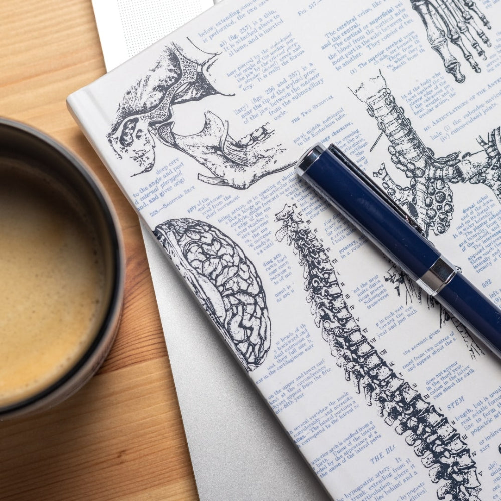 Notebook with anatomical illustrations, a pen, and a cup of coffee on a wooden surface