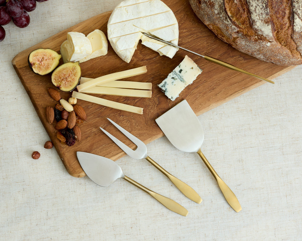 Wooden cutting board with assorted cheeses, figs, nuts, and bread, accompanied by cheese knives on a light background.