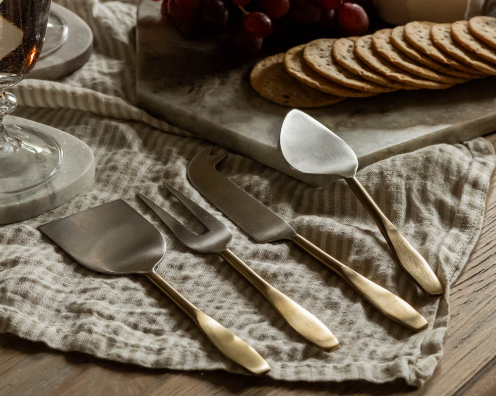 Set of cheese gold cutlery on a textured cloth with a glass and bread slices in the background.
