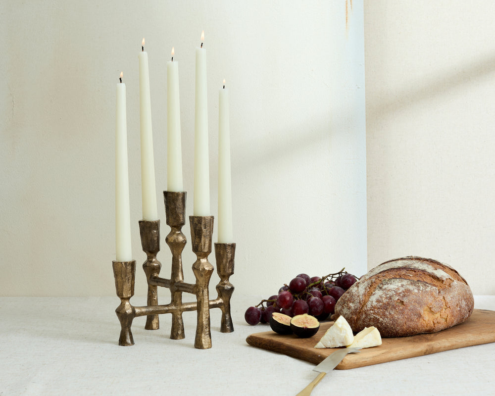 Bronze candle holder with lit candles on a white surface, accompanied by a loaf of bread and fruits.