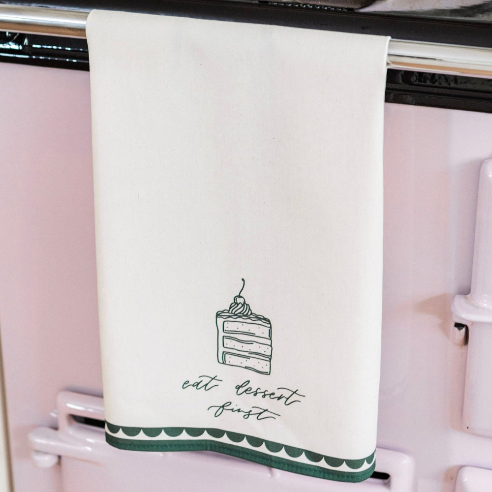 White tea towel with a cake illustration and 'eat dessert first' text hanging on a pink refrigerator.