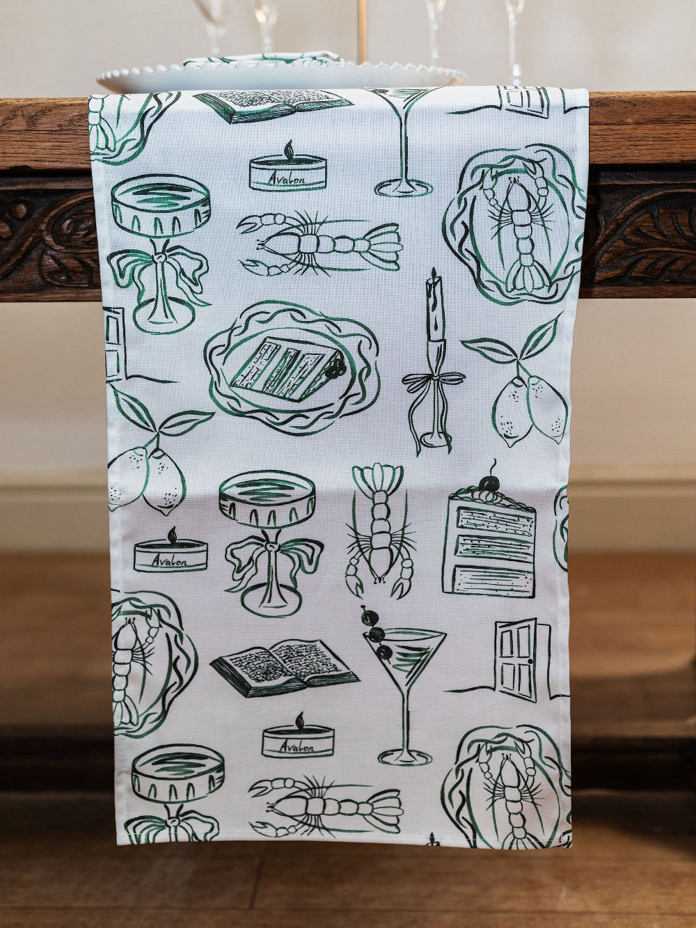 Table runner with art illustrations of martini glasses, champagne glasses, lobsters, books on a white background, hanging on a wooden table.
