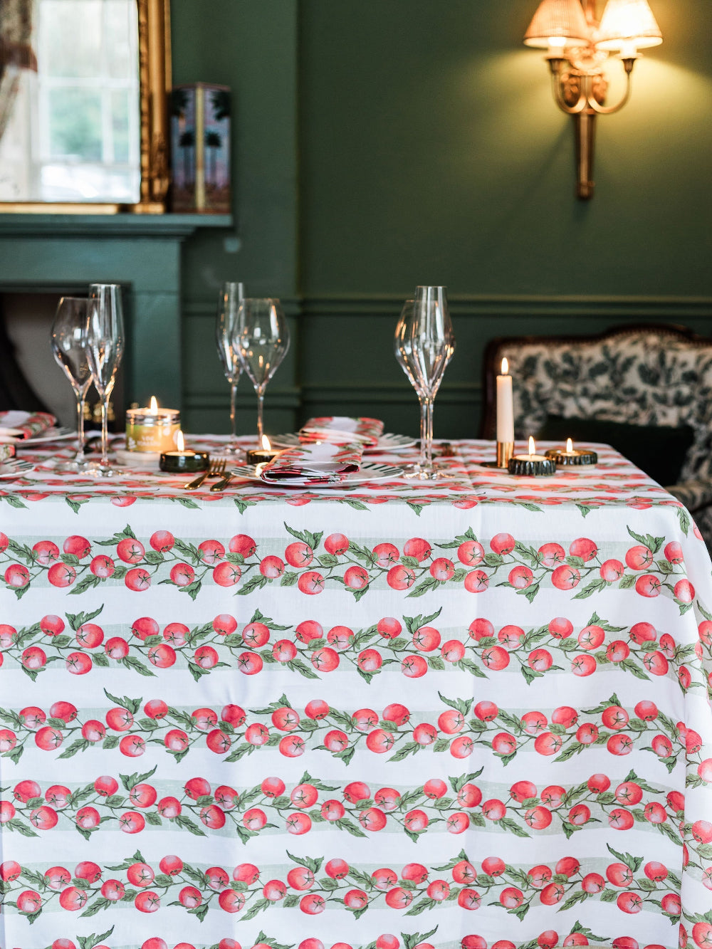 Dining table set with a tomato-patterned tablecloth in a room with green walls and a fireplace.