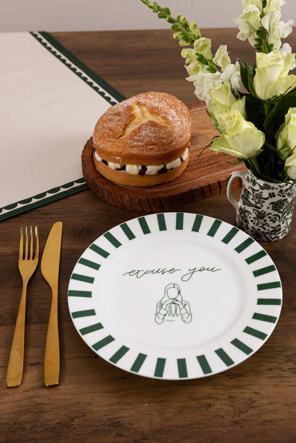 Dessert with a cookie and cream filling on a wooden board, accompanied by a plate with green stripes and a small illustration, a cup of flowers, and cutlery on a wooden table.