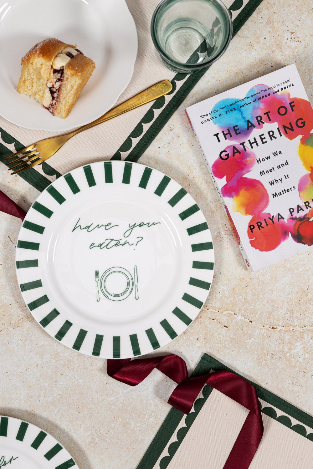 Table setting with 'have you eaten' plate, a glass, a book titled 'The Art of Gathering', and a piece of bread.