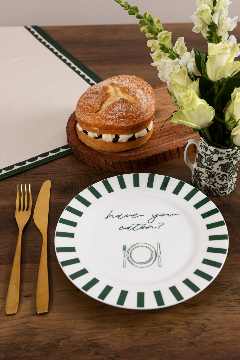 Dessert on a wooden board, flowers in a vase, and a decorative plate with 'have you eaten' text on a wooden table.