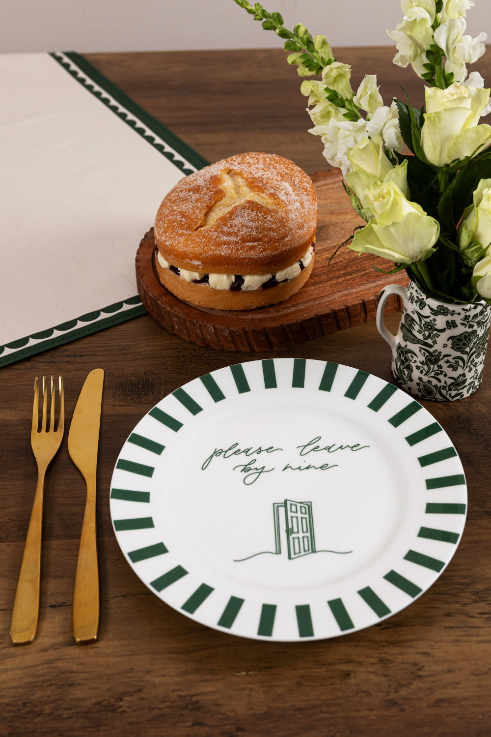 Table setting with a bone china plate, cake, flowers, and cutlery on a wooden surface.