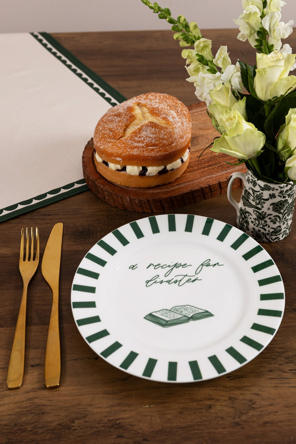 Dessert with a roll and cream on a wooden board, next to a plate with text 'a recipe for disaster' and a cup of flowers on a wooden table. 