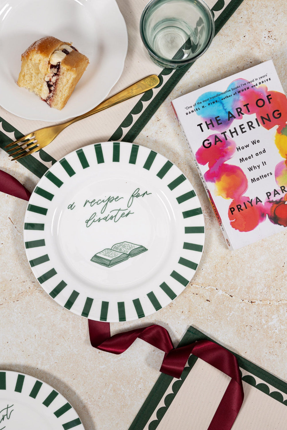 Table setting with a striped plate 'recipe for disaster' text, a book titled 'The Art of Gathering', and a pastry.