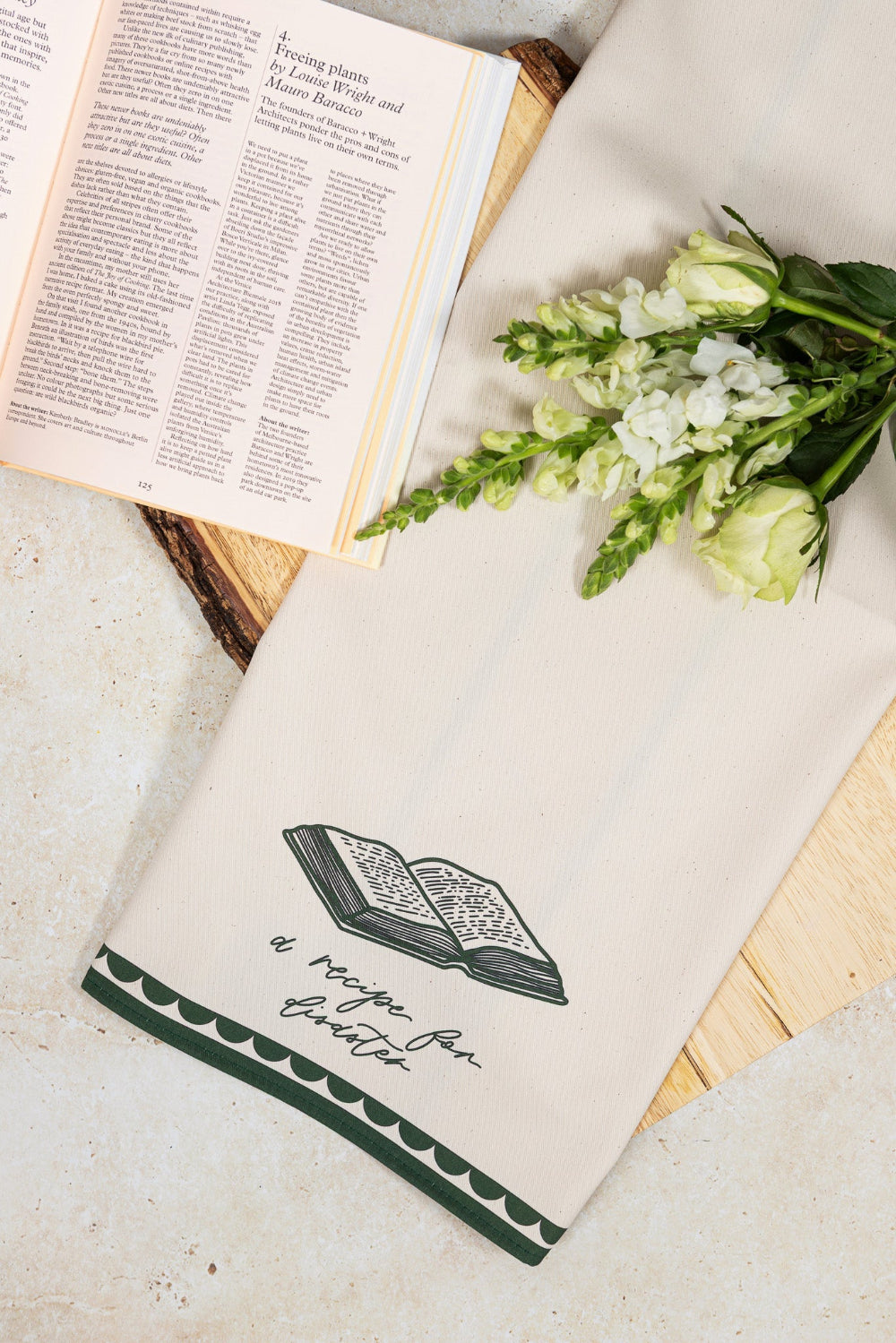 Tea towel with text 'recipe for disaster' placed on a surface with an open book and flowers.