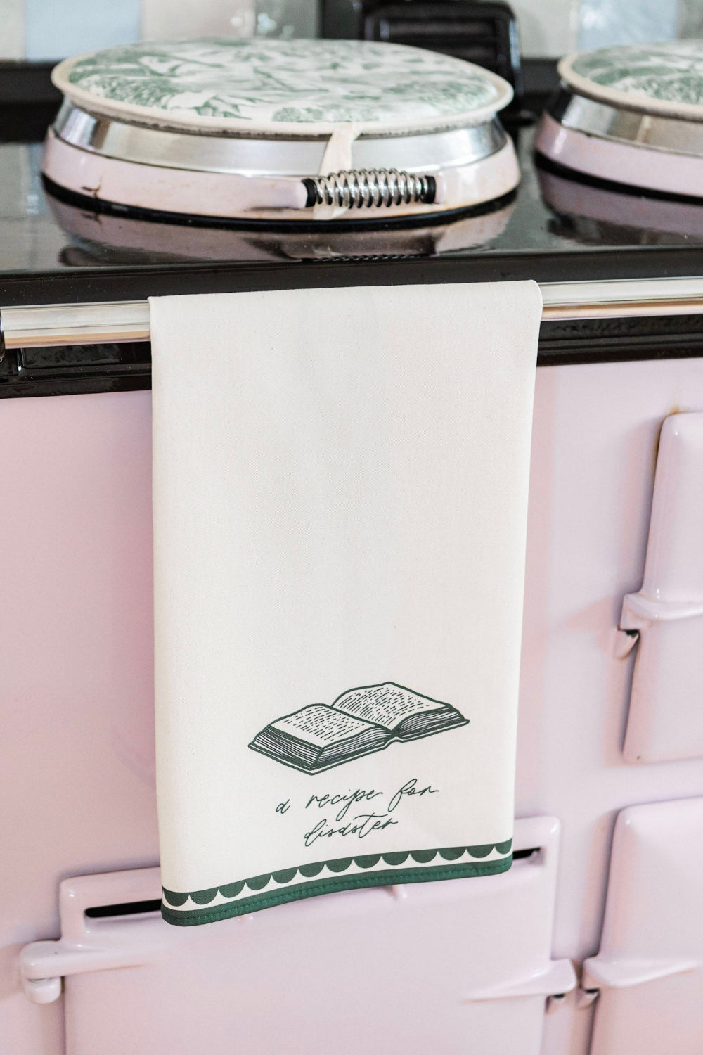 Kitchen tea towel with 'a recipe for disaster' slogan hanging on a pink stove.