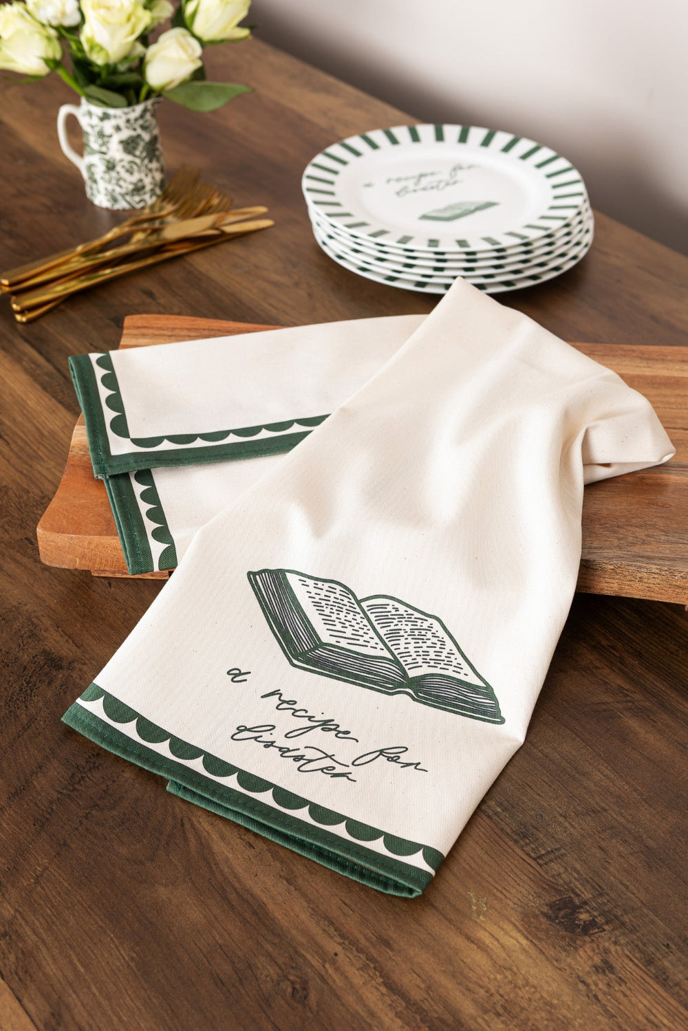 Kitchen towel with 'recipe for disaster' text and on a wooden table with plates and flowers.