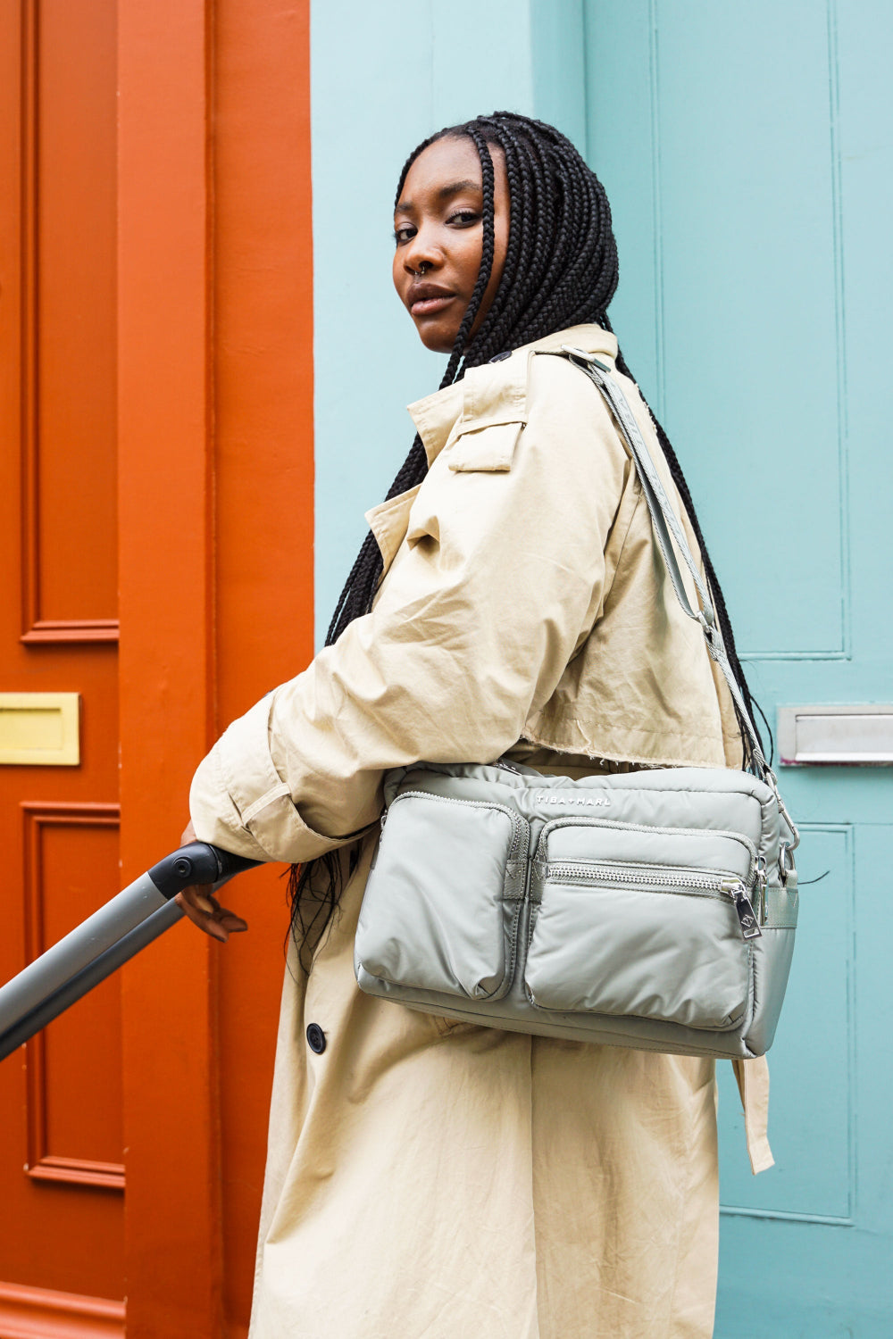 Woman wearing a beige coat with a sage green puffer baby organiser bag against a colourful door background