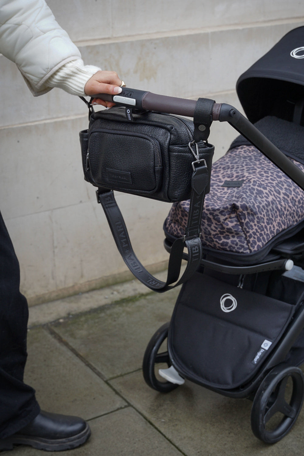 Person holding a black vegan leather bag attached to a stroller on a sidewalk.