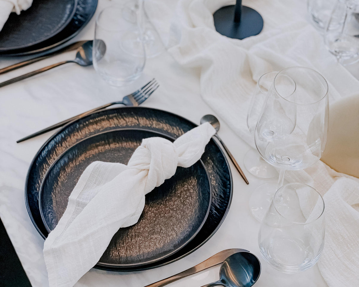 Dining table setting with black plates, white napkins, and clear glasses on a white tablecloth.