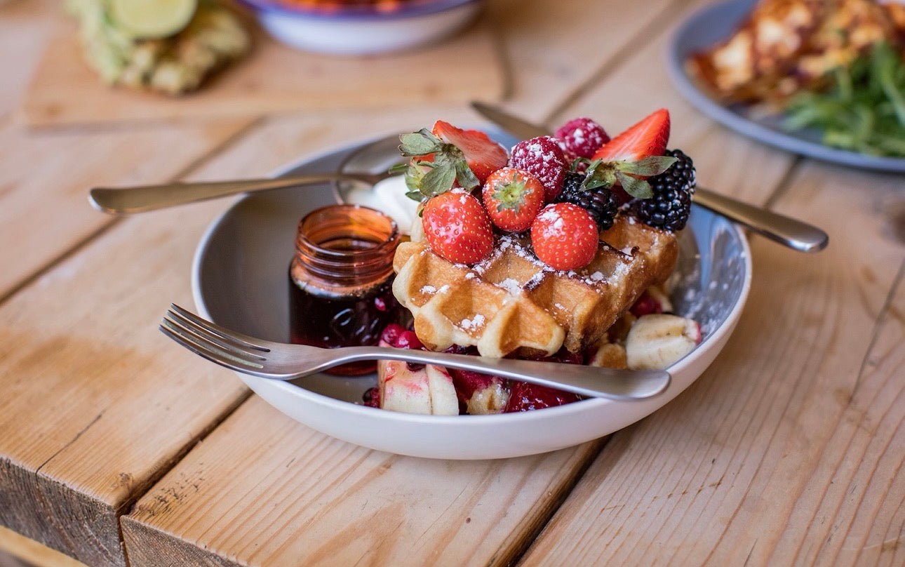 Breakfast Bowl with waffle, honey, fruit, icing sugar and cutlery on a wooden table