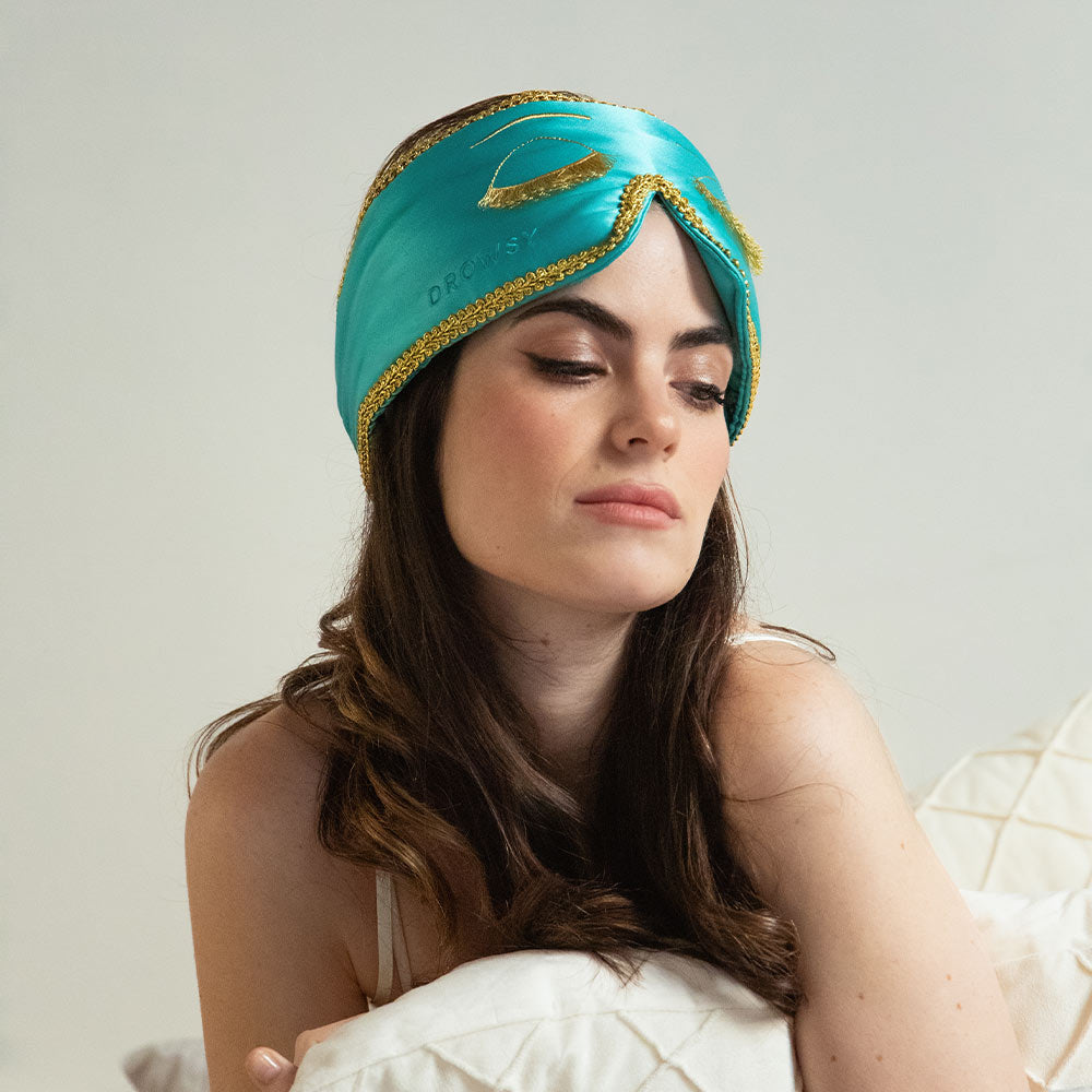 Woman wearing a turquoise Breakfast at Tiffany's inspired sleep eye mask with gold accents with winged Eyeliner on a bed holding a pillow.