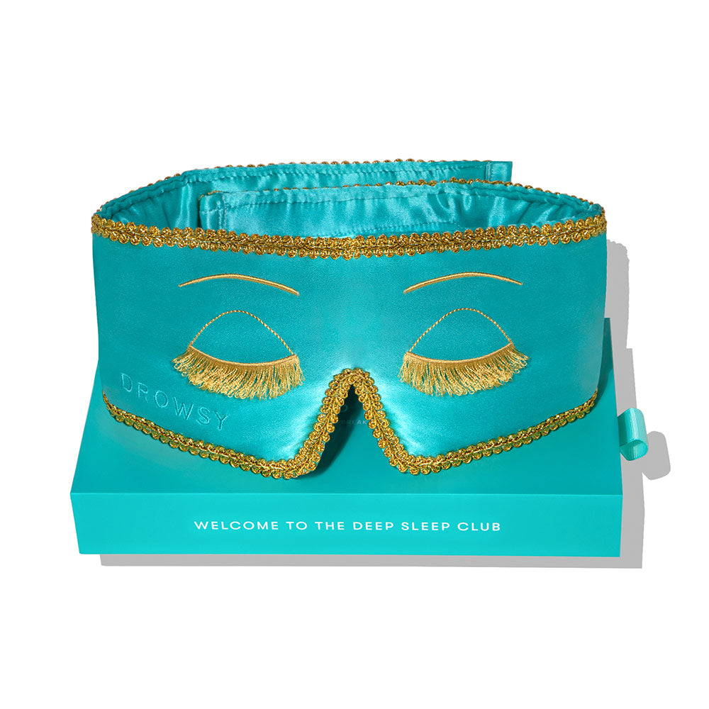 Turquoise sleep mask with gold accents on a white background on branded packaging 'Welcome to the deep sleep club' Breakfast at Tiffany's inspired mask