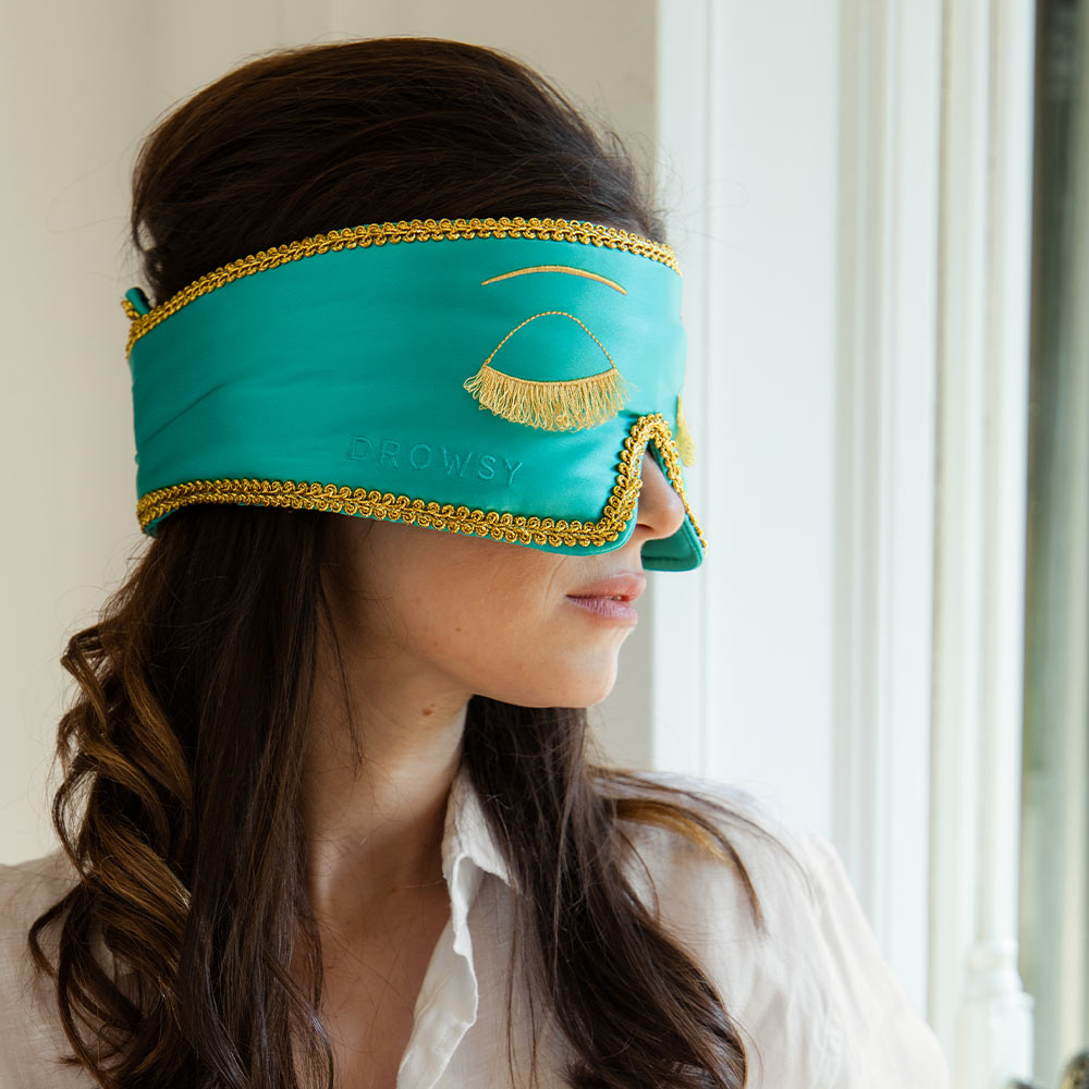Woman wearing a silk Breakfast at Tiffany's inspired sleep eye mask with gold accents facing a window with white netting.