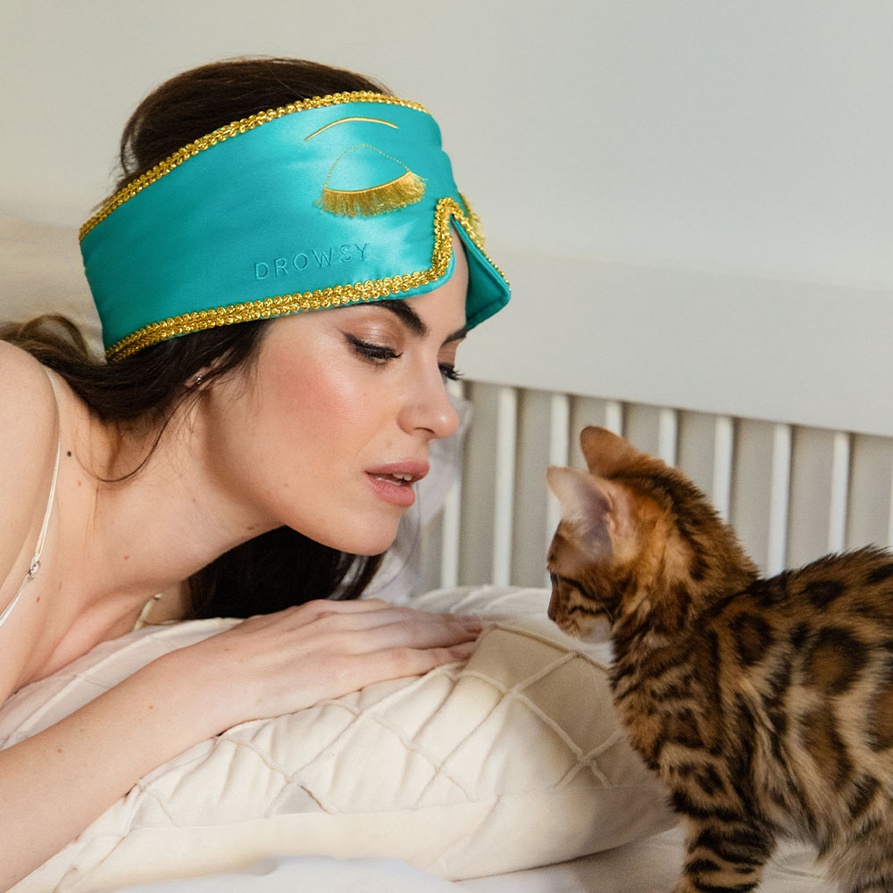 Woman wearing a Breakfast at Tiffany's inspired eye mask with a cat beside her on a bed.