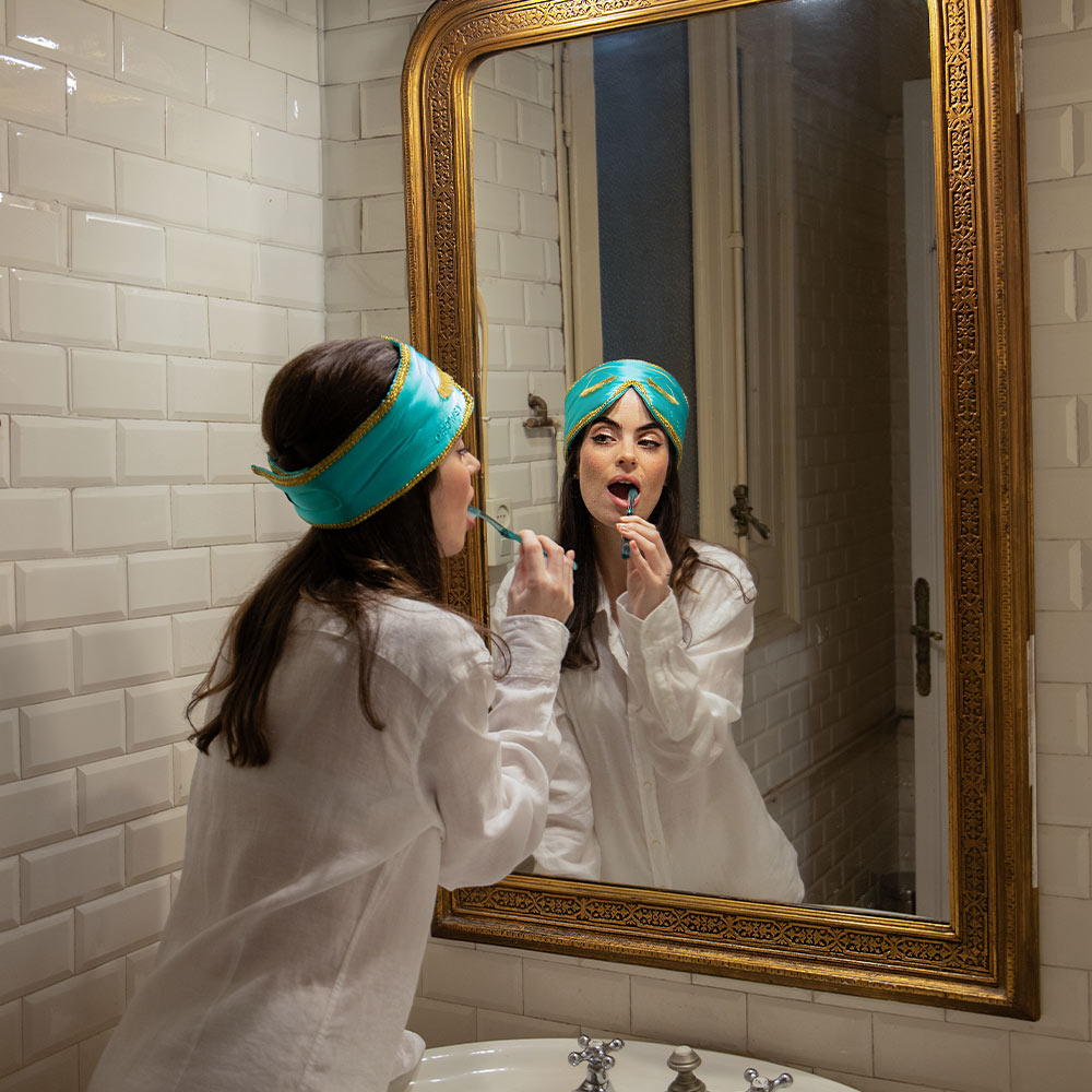 Woman brushing her teeth in front of a large mirror with a gold frame wearing Audrey Hepburn Breakfast at Tiffany's inspired silk eye mask.