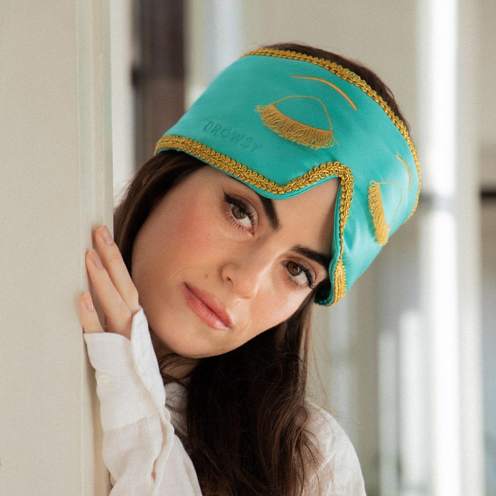 Woman wearing a teal Breakfast at Tiffany's inspired silk eye mask with gold accents looking around a doorframe into a room.