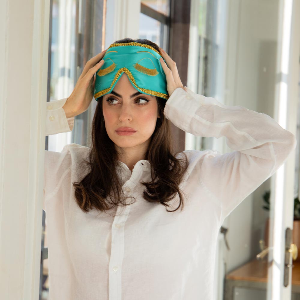 Woman wearing a Breakfast at Tiffany's inspired silk sleep eye mask with gold accents in a light and bright room.