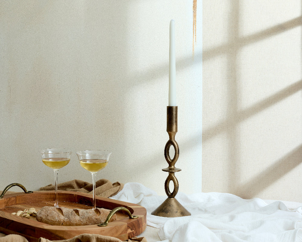 Brass candlestick with a white candle on a wooden tray with glasses of white wine against a neutral background