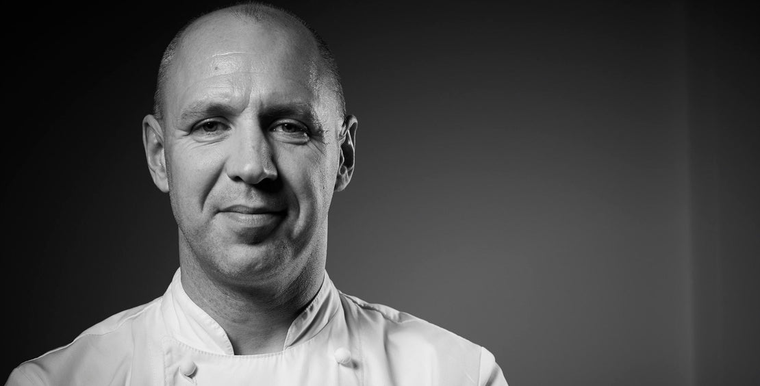 Black and white portrait of a chef wearing a white coat against a dark background