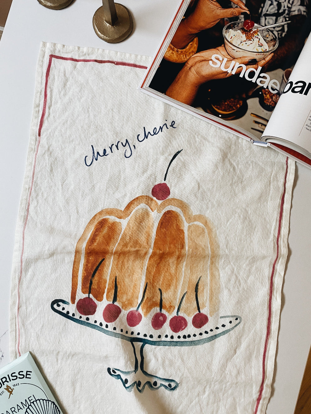 Handmade tea towel with cake illustration and 'Cherry, cherie' text on a table with a magazine.