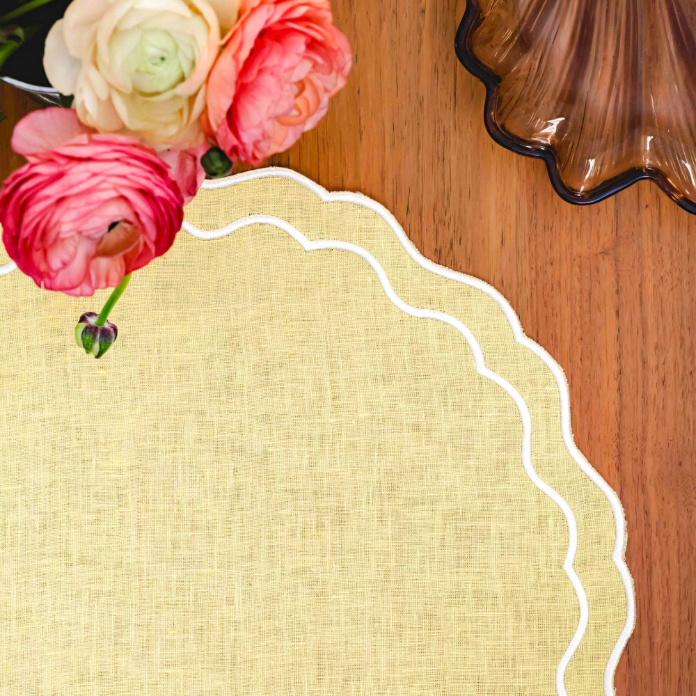 Decorative yellow placemat with scalloped edge on a wooden surface with flowers and a decorative item.