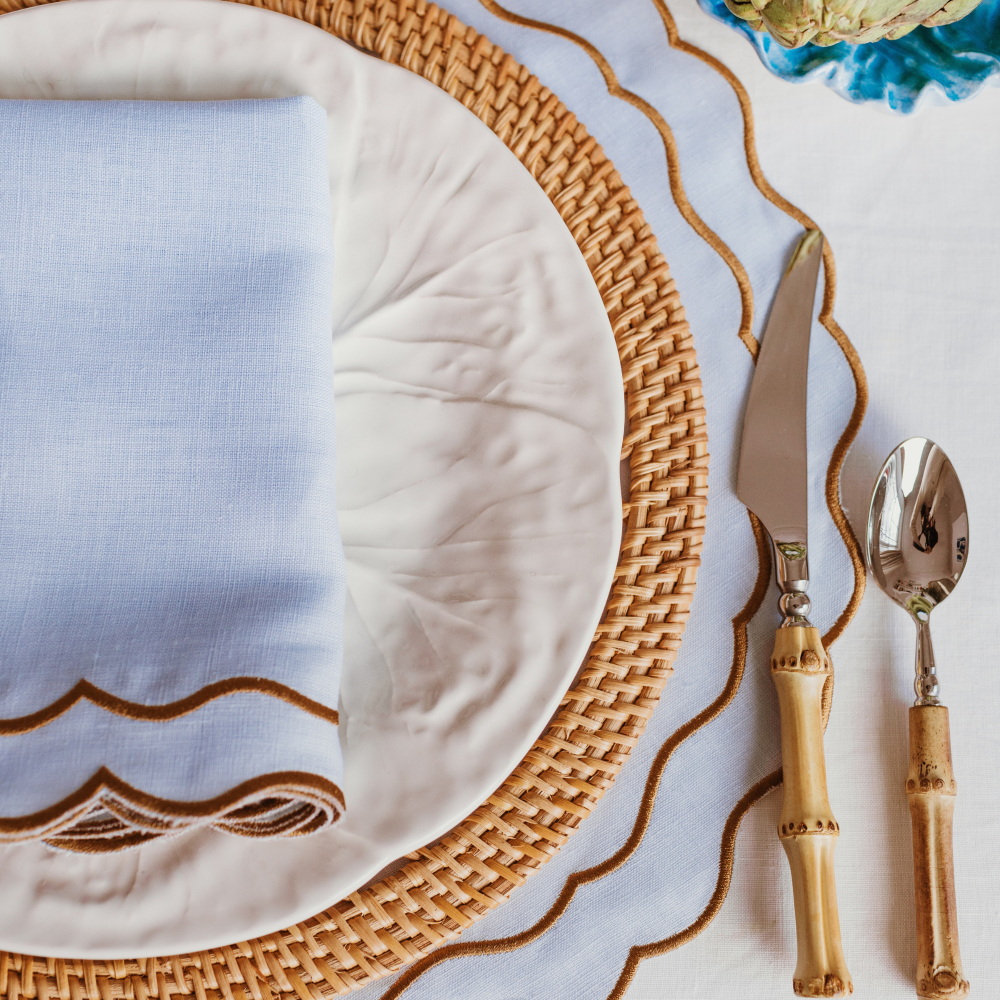 Dining table setting with woven placemat, white plate, and bamboo cutlery on a decorative tablecloth.
