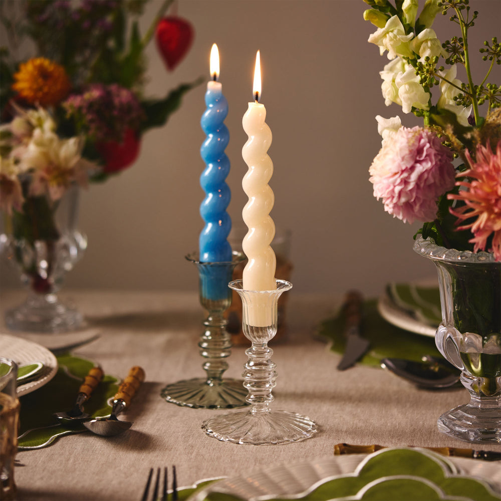 Two twisted candles in glass holders on a table with flowers and cutlery.